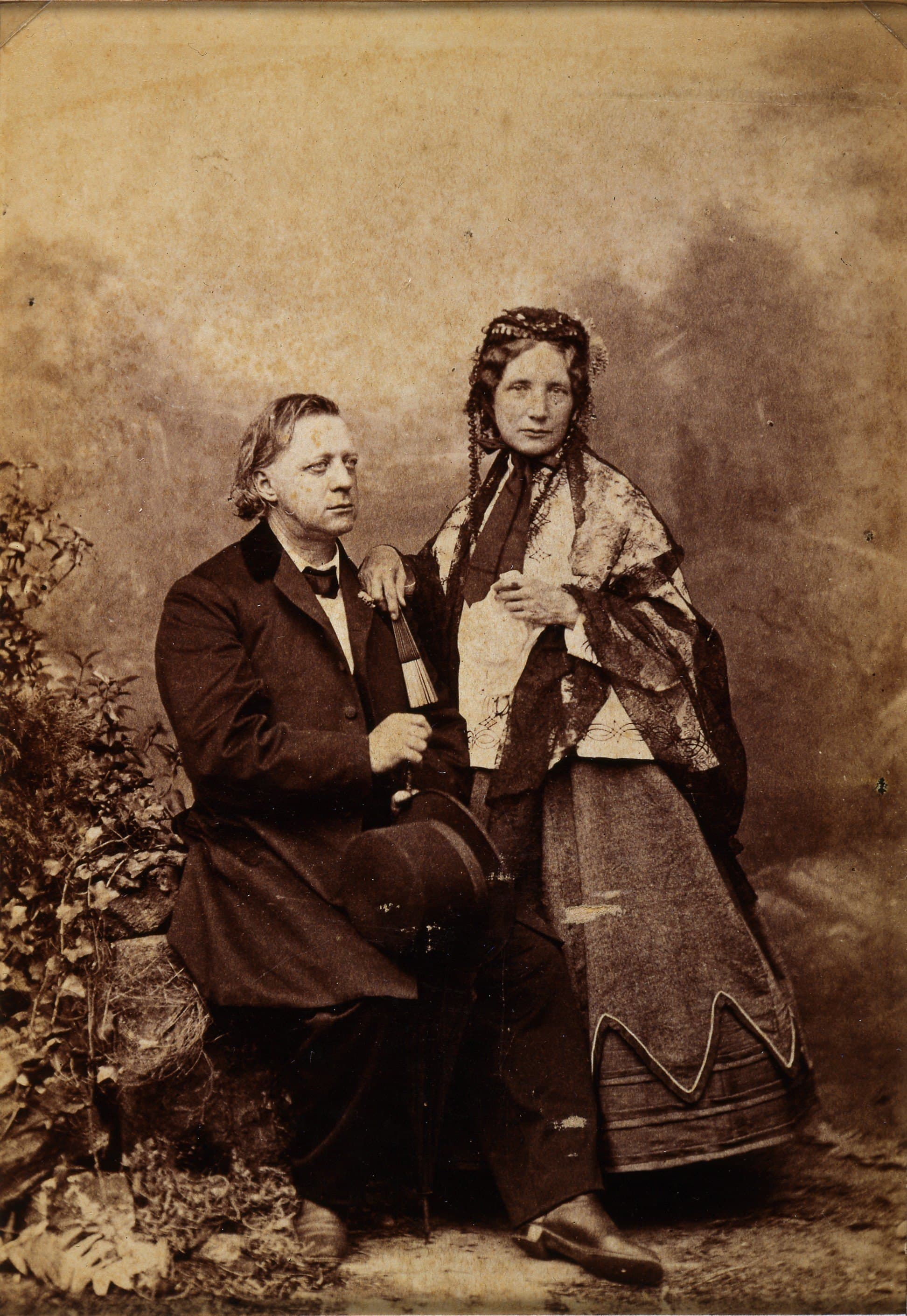 Sepia studio portrait of Harriet Beecher Stowe standing beside her husband, who is seated with a top hat