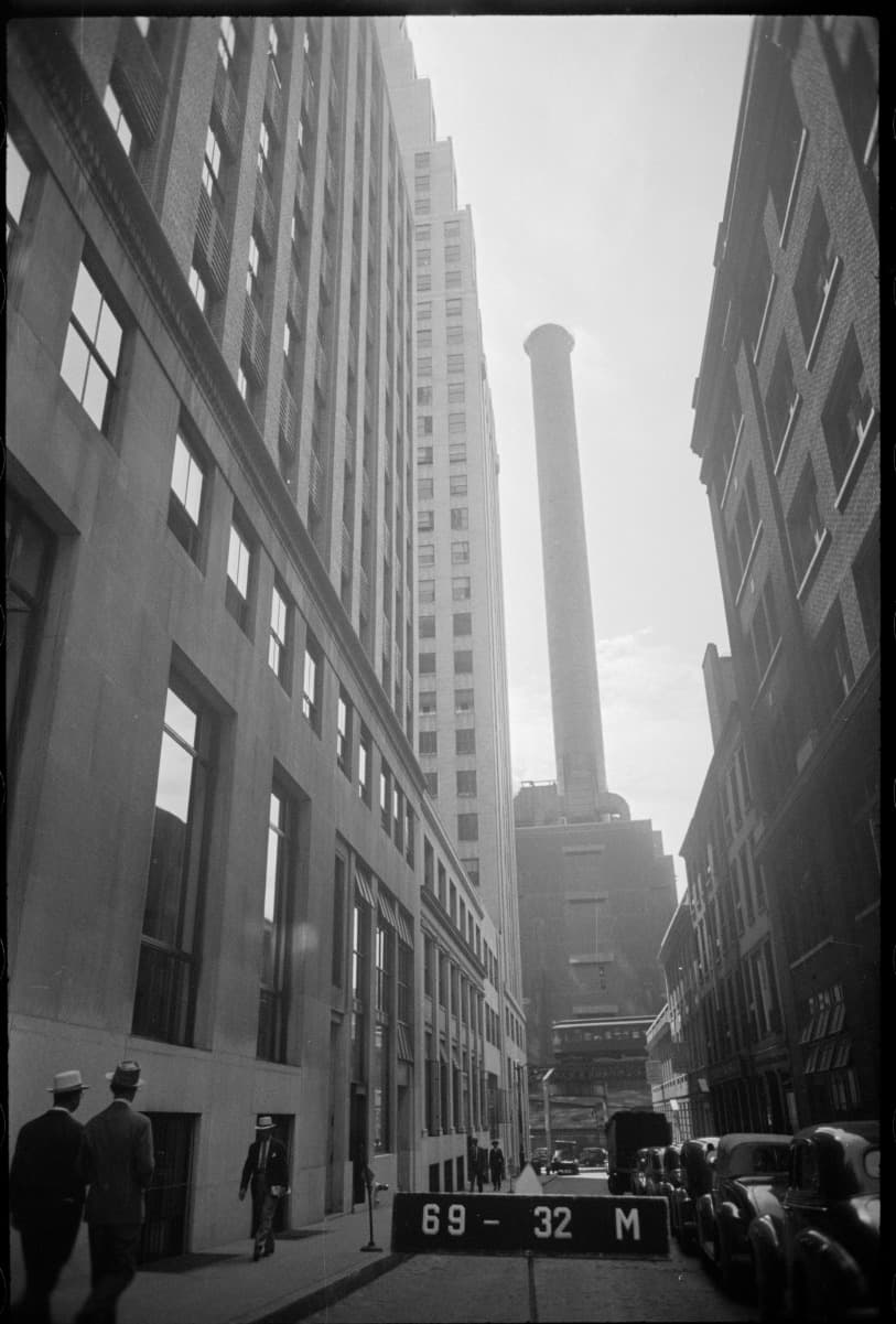 Lower Manhattan street canyon with a tall smokestack and 1940s cars beside 116 John Street