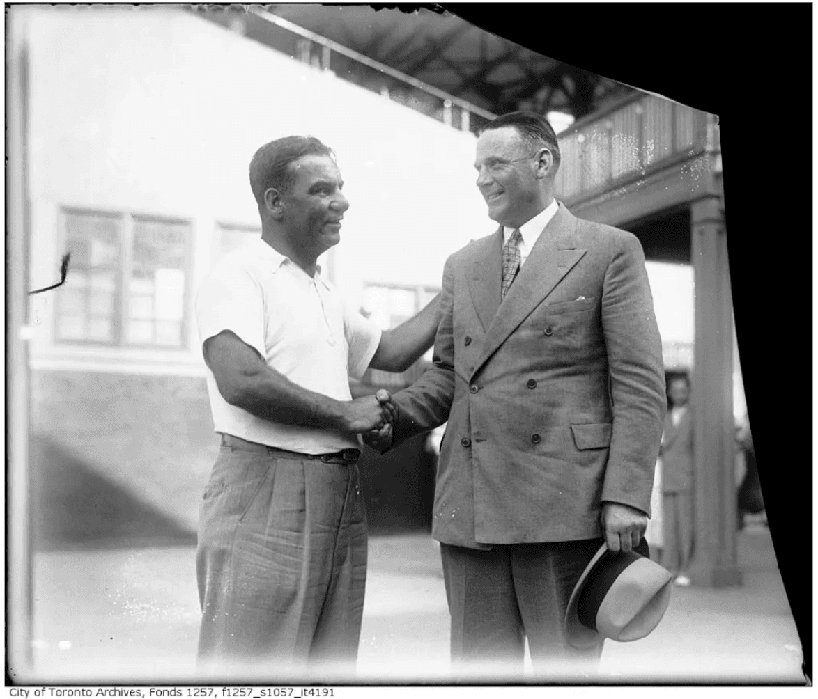 Archival photograph of Norman Red Ryan shaking hands with a man in a suit, holding a hat